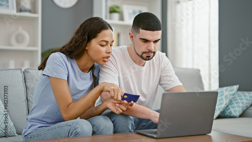 Beautiful couple in love, sitting together on sofa, engrossed in online shopping using a laptop and credit card, enjoying a relaxed lifestyle within their home's living room interior.