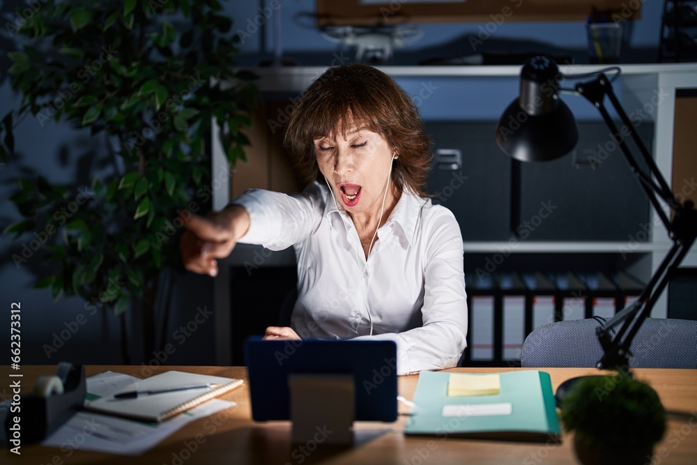 Middle age woman working at the office at night pointing with finger surprised ahead, open mouth amazed expression, something on the front