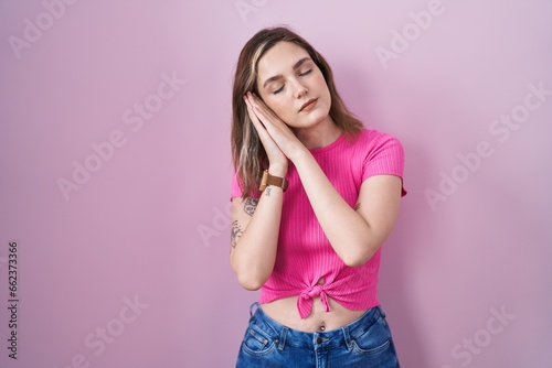 Blonde caucasian woman standing over pink background sleeping tired dreaming and posing with hands together while smiling with closed eyes.