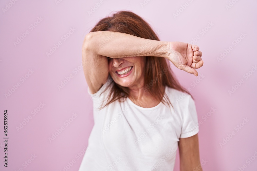 Middle age woman standing over pink background covering eyes with arm smiling cheerful and funny. blind concept. Middle age woman standing over pink background covering eyes with arm smiling cheerful and funny. blind concept.