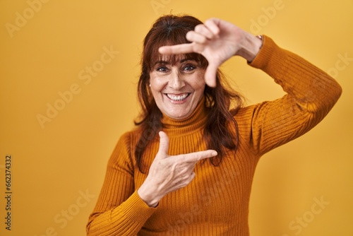 Middle age hispanic woman standing over yellow background smiling making frame with hands and fingers with happy face. creativity and photography concept.