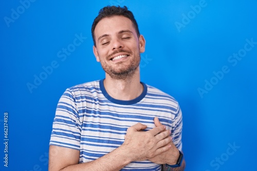 Young hispanic man standing over blue background smiling with hands on chest with closed eyes and grateful gesture on face. health concept.