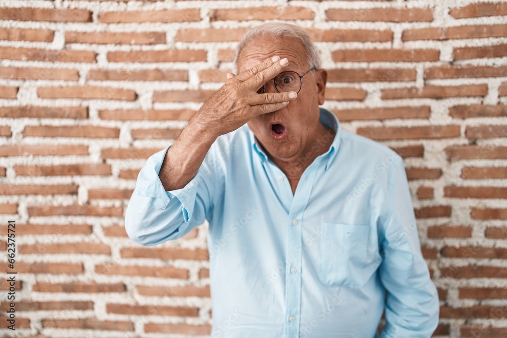Senior man with grey hair standing over bricks wall peeking in shock covering face and eyes with hand, looking through fingers with embarrassed expression. Senior man with grey hair standing over bricks wall peeking in shock covering face and eyes with hand, looking through fingers with embarrassed expression.