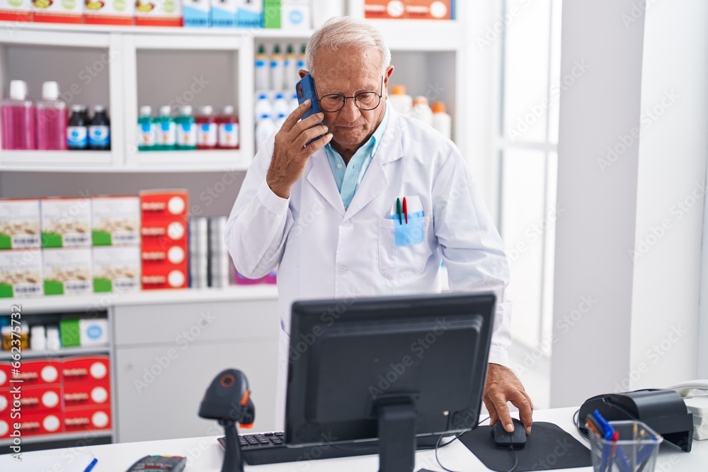 Senior grey-haired man pharmacist talking on smartphone using computer at pharmacy