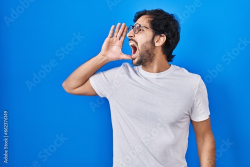 Handsome latin man standing over blue background shouting and screaming loud to side with hand on mouth. communication concept.