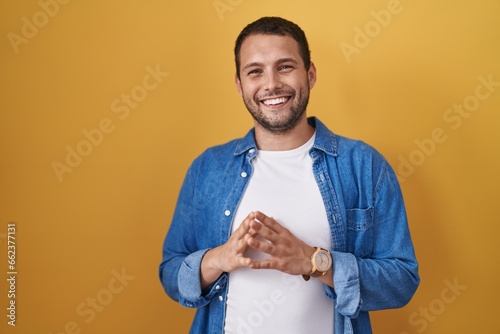 Canvas Print Hispanic man standing over yellow background hands together and fingers crossed smiling relaxed and cheerful