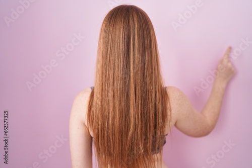 Redhead woman wearing lingerie over pink background posing backwards pointing ahead with finger hand