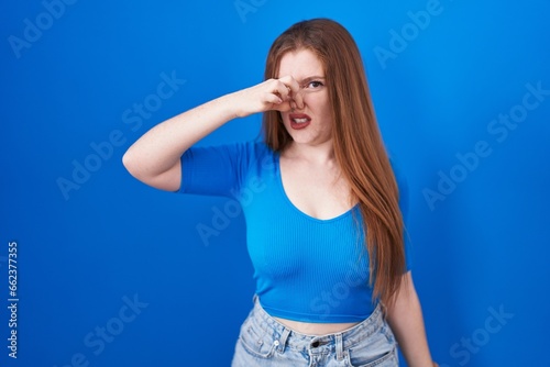 Redhead woman standing over blue background smelling something stinky and disgusting, intolerable smell, holding breath with fingers on nose. bad smell