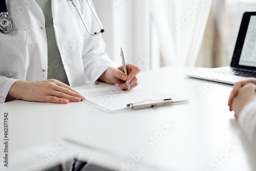 Fototapeta Naklejka Na Ścianę i Meble -  Doctor and patient sitting near each other at the white desk in clinic. Female physician is listening filling up a records form. Medicine concept
