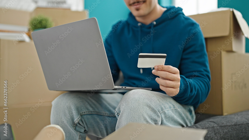 Young hispanic man shopping with laptop and credit card sitting on sofa at new home Young hispanic man shopping with laptop and credit card sitting on sofa at new home