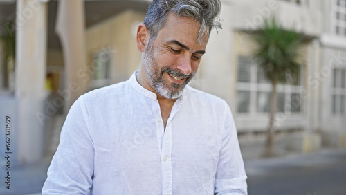 Young grey-haired hispanic man, confidently smiling, standing, basking in sunny street ambiance, a cool look in his eyes, exuding positivity.