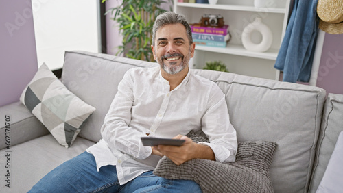 Confident young hispanic man with grey hair, joyfully using touchpad on comfy sofa in his indoor apartment living room, expressing happiness through his handsome bearded smile