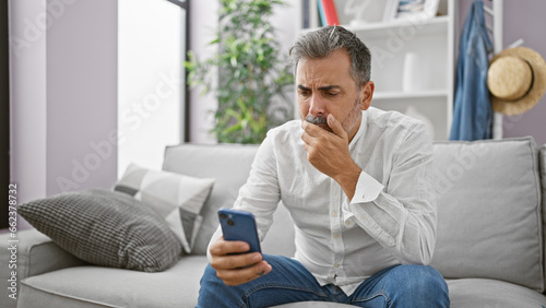 Upset young hispanic man, sporting grey hair and a beard, engrossed in a serious smartphone ordeal while comfortably nestled on his living room sofa at home