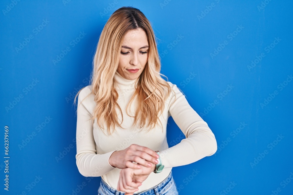 Young caucasian woman standing over blue background checking the time on wrist watch, relaxed and confident Young caucasian woman standing over blue background checking the time on wrist watch, relaxed and confident