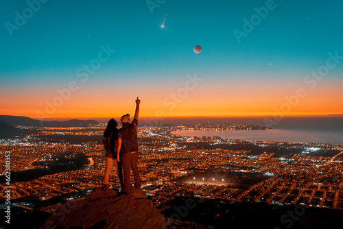 silhouettes of a couple standing on the top of the hill looking at starry night with Moon and shooting star in the sky over the city light 