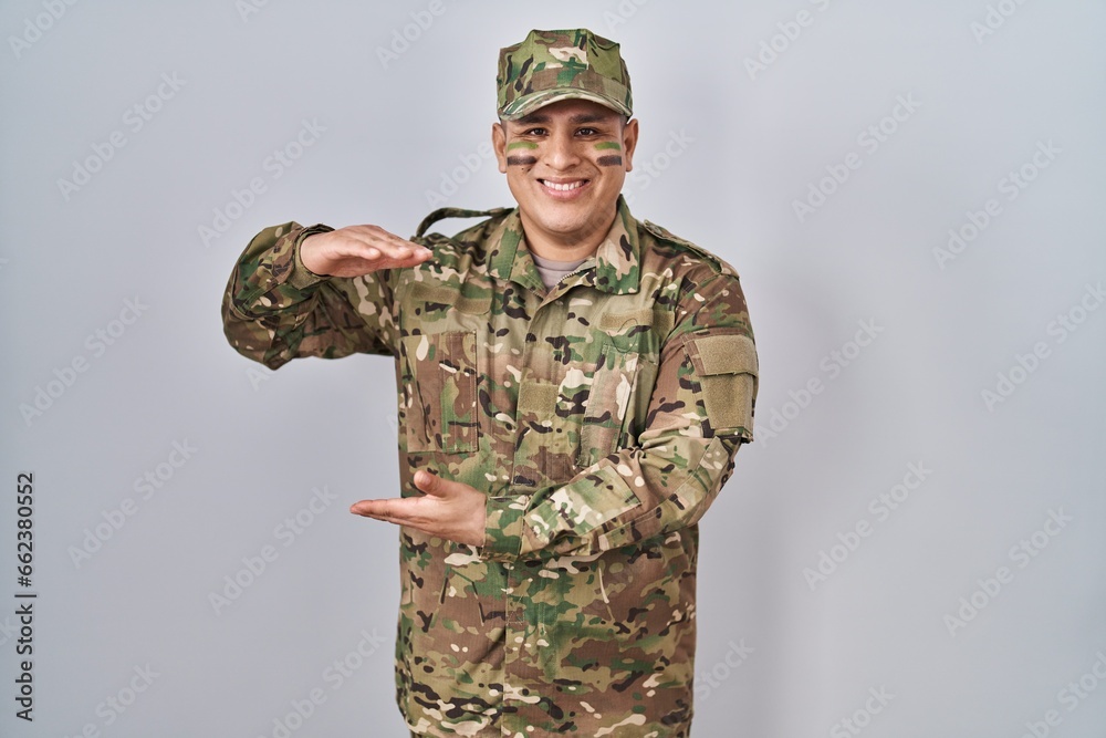 Hispanic young man wearing camouflage army uniform gesturing with hands showing big and large size sign, measure symbol. smiling looking at the camera. measuring concept. Hispanic young man wearing camouflage army uniform gesturing with hands showing big and large size sign, measure symbol. smiling looking at the camera. measuring concept.