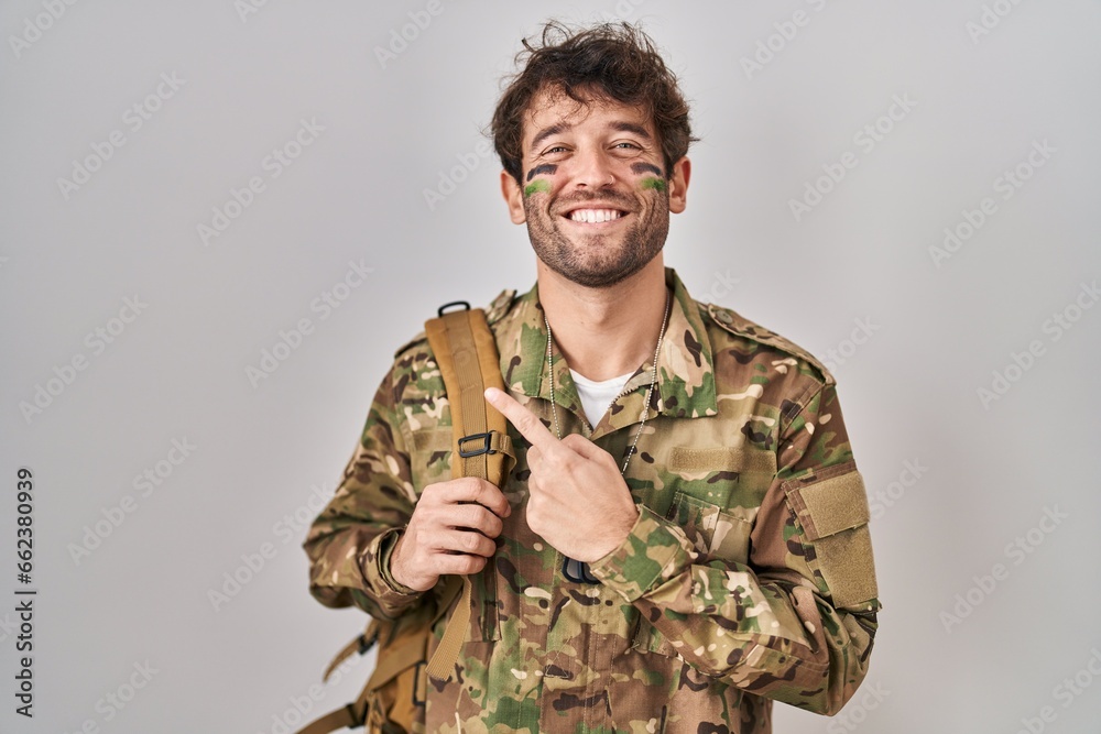 Hispanic young man wearing camouflage army uniform cheerful with a smile on face pointing with hand and finger up to the side with happy and natural expression