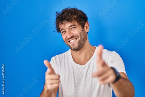 Hispanic young man standing over blue background pointing fingers to camera with happy and funny face. good energy and vibes.