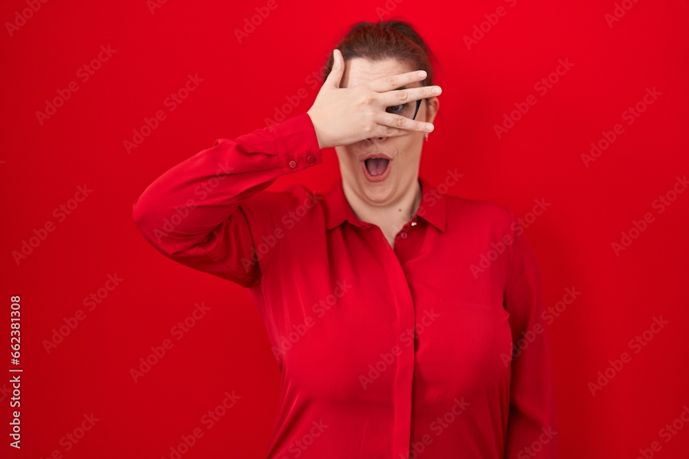 Young hispanic woman with red hair standing over red background peeking in shock covering face and eyes with hand, looking through fingers with embarrassed expression.