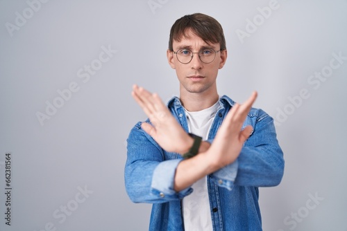 Caucasian blond man standing wearing glasses rejection expression crossing arms and palms doing negative sign, angry face