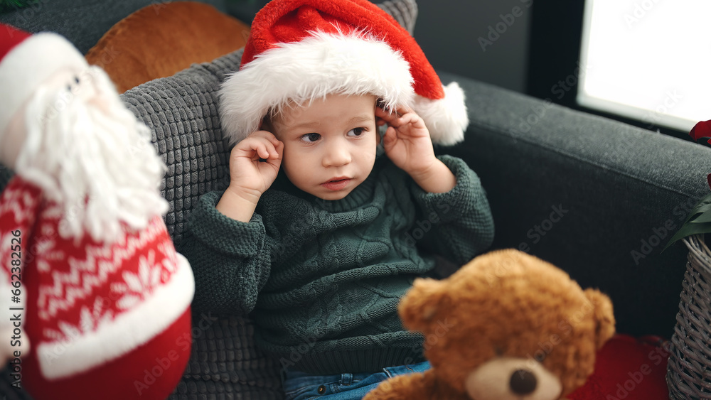 Adorable blond toddler sitting on sofa wearing christmas hat at home