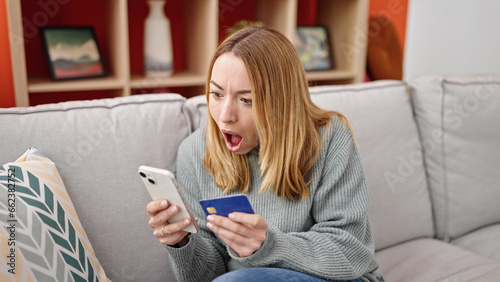 Young blonde woman shopping with smartphone and credit card with surprise expression at home