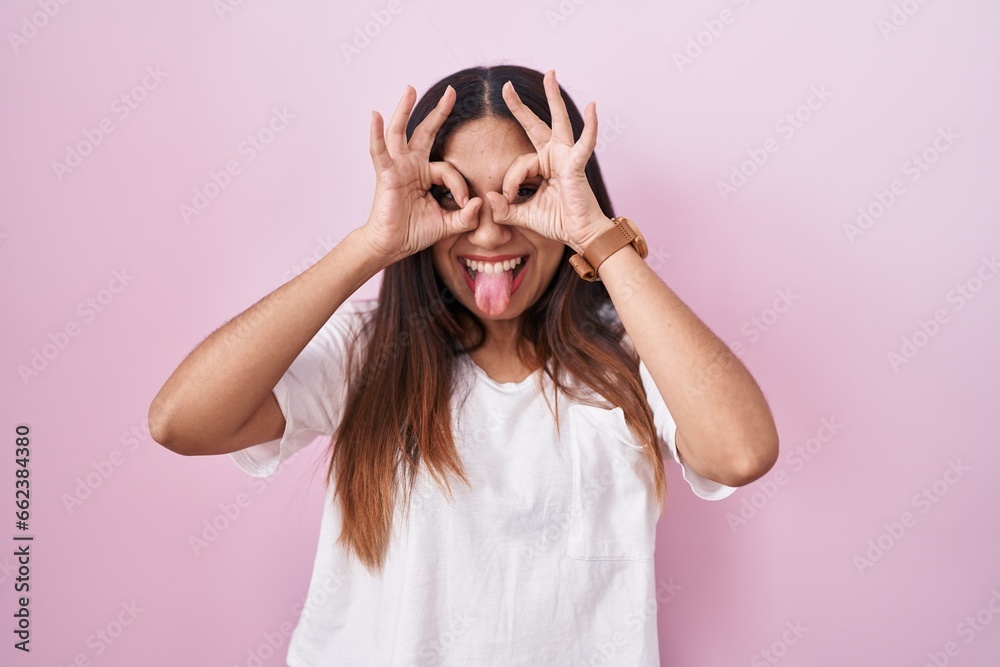 Young arab woman standing over pink background doing ok gesture like binoculars sticking tongue out, eyes looking through fingers. crazy expression. Young arab woman standing over pink background doing ok gesture like binoculars sticking tongue out, eyes looking through fingers. crazy expression.