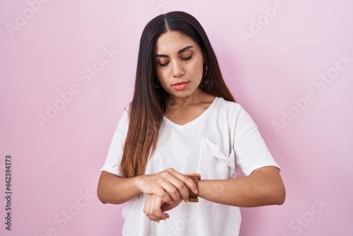 Young arab woman standing over pink background checking the time on wrist watch  relaxed and confident