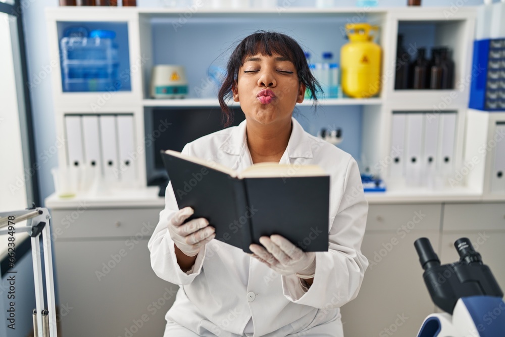 Hispanic woman working at scientist laboratory reading a book looking at the camera blowing a kiss being lovely and sexy. love expression. Hispanic woman working at scientist laboratory reading a book looking at the camera blowing a kiss being lovely and sexy. love expression.