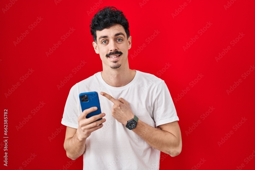 Hispanic man using smartphone over red background cheerful with a smile on face pointing with hand and finger up to the side with happy and natural expression Hispanic man using smartphone over red background cheerful with a smile on face pointing with hand and finger up to the side with happy and natural expression