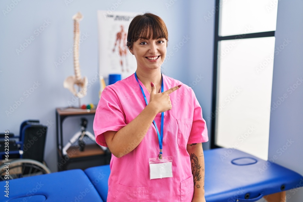 Young brunette woman working at rehabilitation clinic cheerful with a smile of face pointing with hand and finger up to the side with happy and natural expression on face