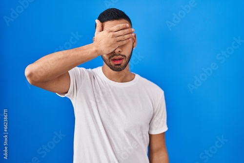 Young hispanic man standing over blue background covering eyes with hand, looking serious and sad. sightless, hiding and rejection concept