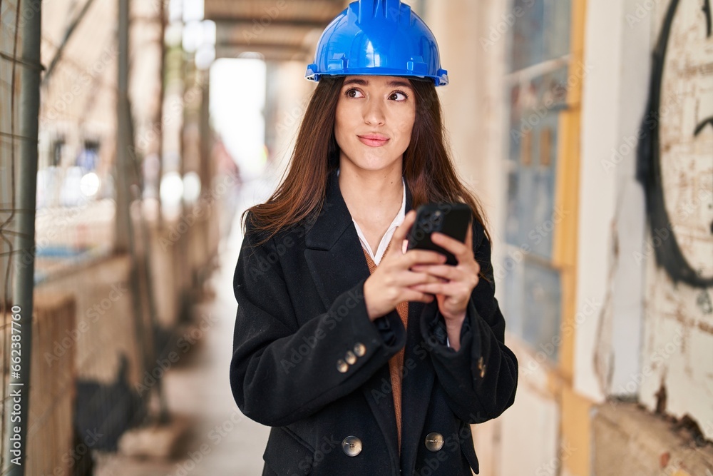 Young beautiful hispanic woman architect using smartphone at street