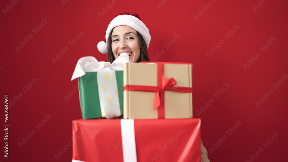 Beautiful hispanic woman wearing christmas hat holding gifts over isolated red background Beautiful hispanic woman wearing christmas hat holding gifts over isolated red background