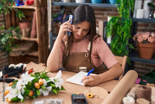 Young beautiful hispanic woman florist talking on smartphone writing on notebook at flower shop