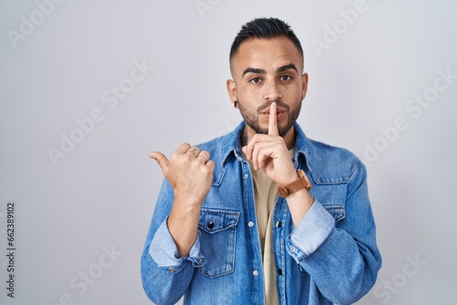 Young hispanic man standing over isolated background asking to be quiet with finger on lips pointing with hand to the side. silence and secret concept.