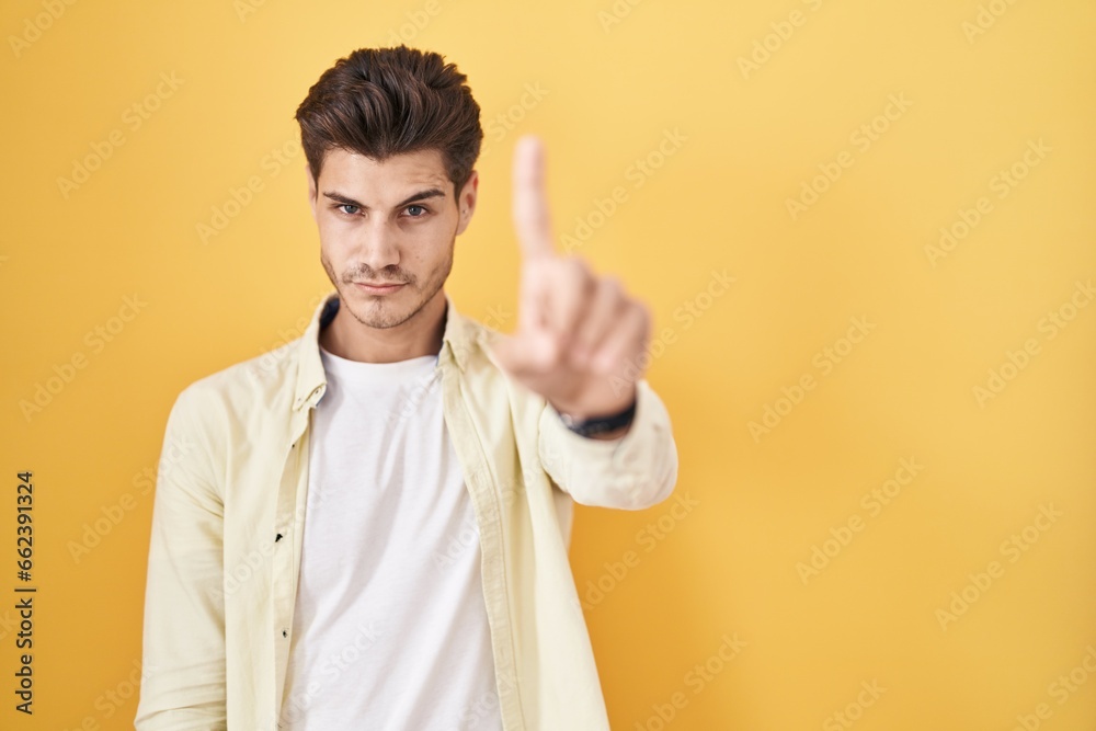 Young hispanic man standing over yellow background pointing with finger up and angry expression, showing no gesture Young hispanic man standing over yellow background pointing with finger up and angry expression, showing no gesture