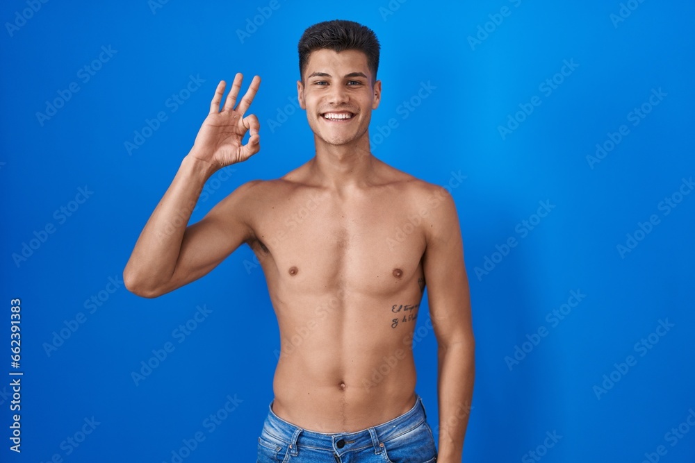 Young hispanic man standing shirtless over blue background smiling positive doing ok sign with hand and fingers. successful expression.