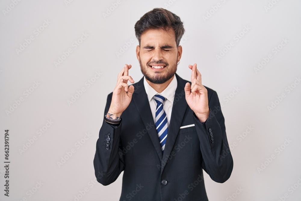 Young hispanic man with tattoos wearing business suit and tie gesturing finger crossed smiling with hope and eyes closed. luck and superstitious concept. Young hispanic man with tattoos wearing business suit and tie gesturing finger crossed smiling with hope and eyes closed. luck and superstitious concept.