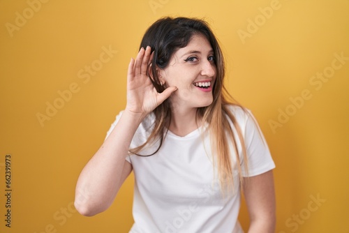 Young brunette woman standing over yellow background smiling with hand over ear listening an hearing to rumor or gossip. deafness concept. © Krakenimages.com