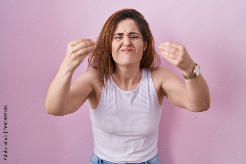 Brunette woman standing over pink background angry and mad raising fists frustrated and furious while shouting with anger. rage and aggressive concept. Brunette woman standing over pink background angry and mad raising fists frustrated and furious while shouting with anger. rage and aggressive concept.