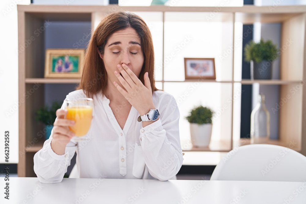 Brunette woman drinking glass of orange juice bored yawning tired covering mouth with hand. restless and sleepiness. Brunette woman drinking glass of orange juice bored yawning tired covering mouth with hand. restless and sleepiness.