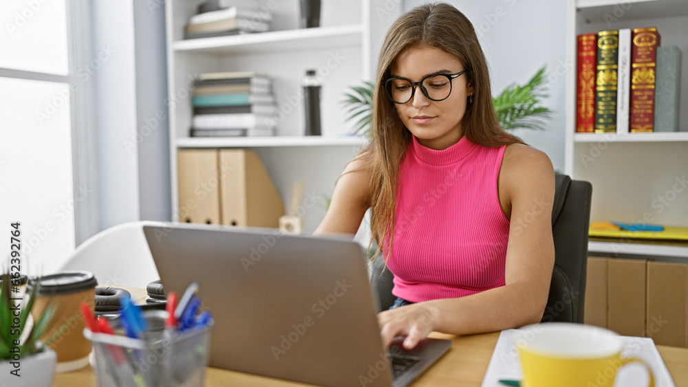 Serious yet relaxed young hispanic woman calmly concentrating on work, beautifying the office with her elegant presence as she skillfully manages her business online on a laptop.