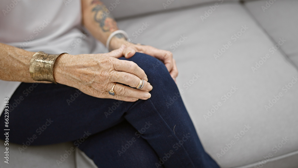 Elderly woman suffering tremendous knee pain, adult, resting despondently in the comfort of her living room sofa, glancing at her injured leg