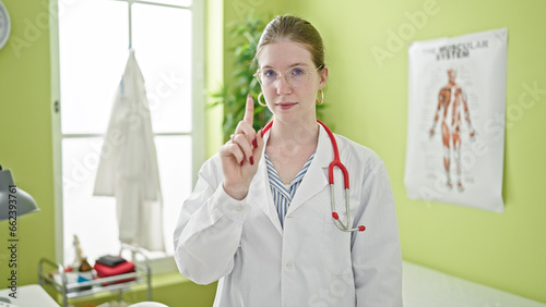 Young blonde woman doctor standing with serious expression saying no with finger at clinic
