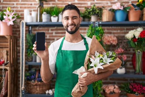 Hispanic young man working at florist shop showing smartphone screen winking looking at the camera with sexy expression, cheerful and happy face.