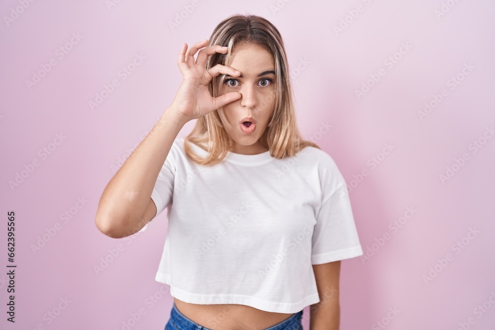 Young blonde woman standing over pink background doing ok gesture shocked with surprised face, eye looking through fingers. unbelieving expression. Young blonde woman standing over pink background doing ok gesture shocked with surprised face, eye looking through fingers. unbelieving expression.