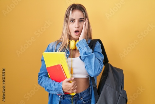 Young blonde woman wearing student backpack and holding books afraid and shocked, surprise and amazed expression with hands on face