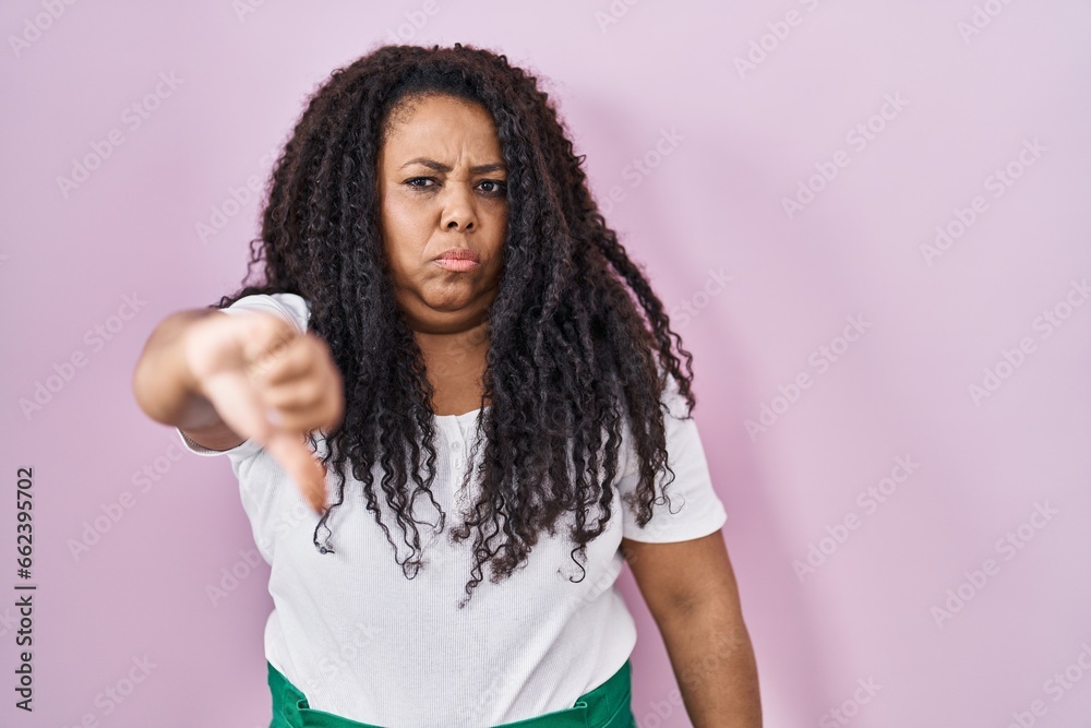 Plus size hispanic woman standing over pink background looking unhappy and angry showing rejection and negative with thumbs down gesture. bad expression. Plus size hispanic woman standing over pink background looking unhappy and angry showing rejection and negative with thumbs down gesture. bad expression.