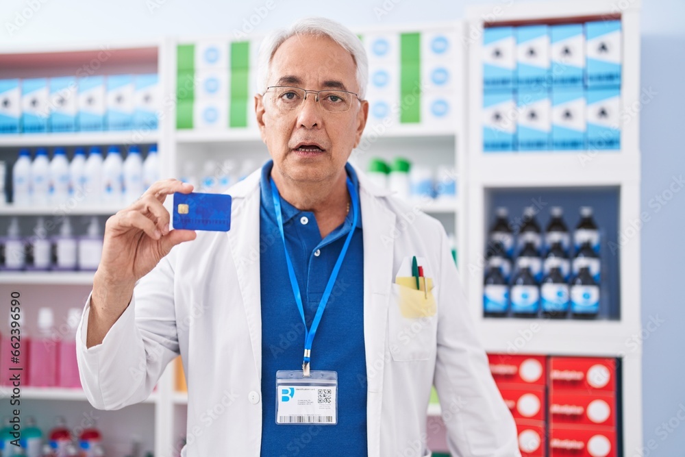 Middle age man with grey hair working at pharmacy drugstore holding credit card scared and amazed with open mouth for surprise, disbelief face